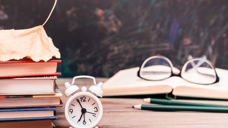 Back to school background with books, clock, fallen leaf, open book and glasses on a wooden table over blackboard